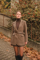 a lady in a brown mock neck sweater, a brown coat with a belt and a pair of brown shorts